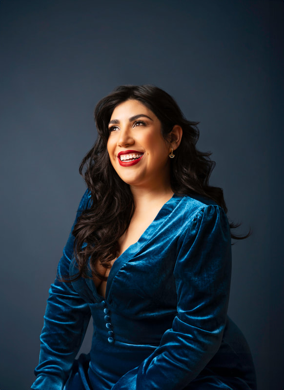 Mexican soprano Judy Yannini is a woman with long dark hair. She is wearing bright red lipstick and a long-sleeved deep teal colored velvet dress.