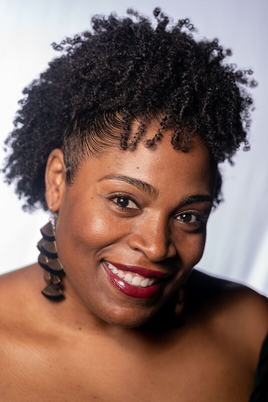 Dr. Monique Spells is a black woman with short, natural hair. She is pictured from the shoulders up and her clothing is not visible. She wears large fan-shaped earrings.